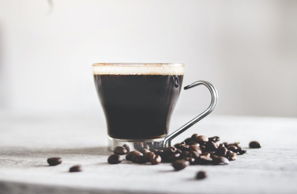 Aromatic black coffee in glass cup with metal handle and scattered coffee beans on light wooden table against blurred background