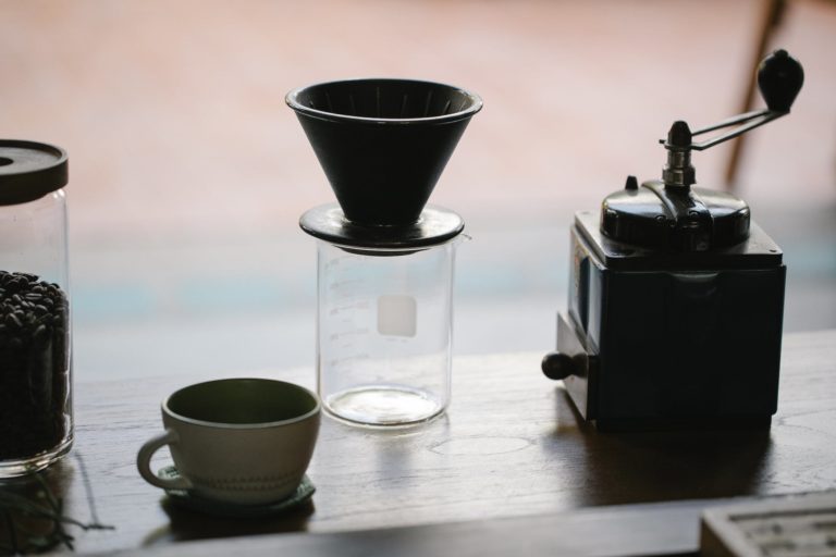 From above of manual coffee grinder with pour over coffeemaker placed in wooden table with ceramic cup and glass jug with roasted beans in morning